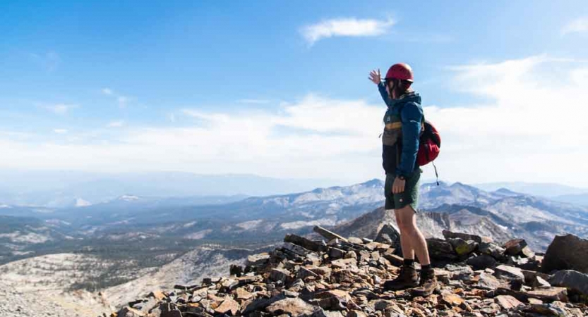 a person wearing a helmet stands on rocky terrain high above a mountainous landscape